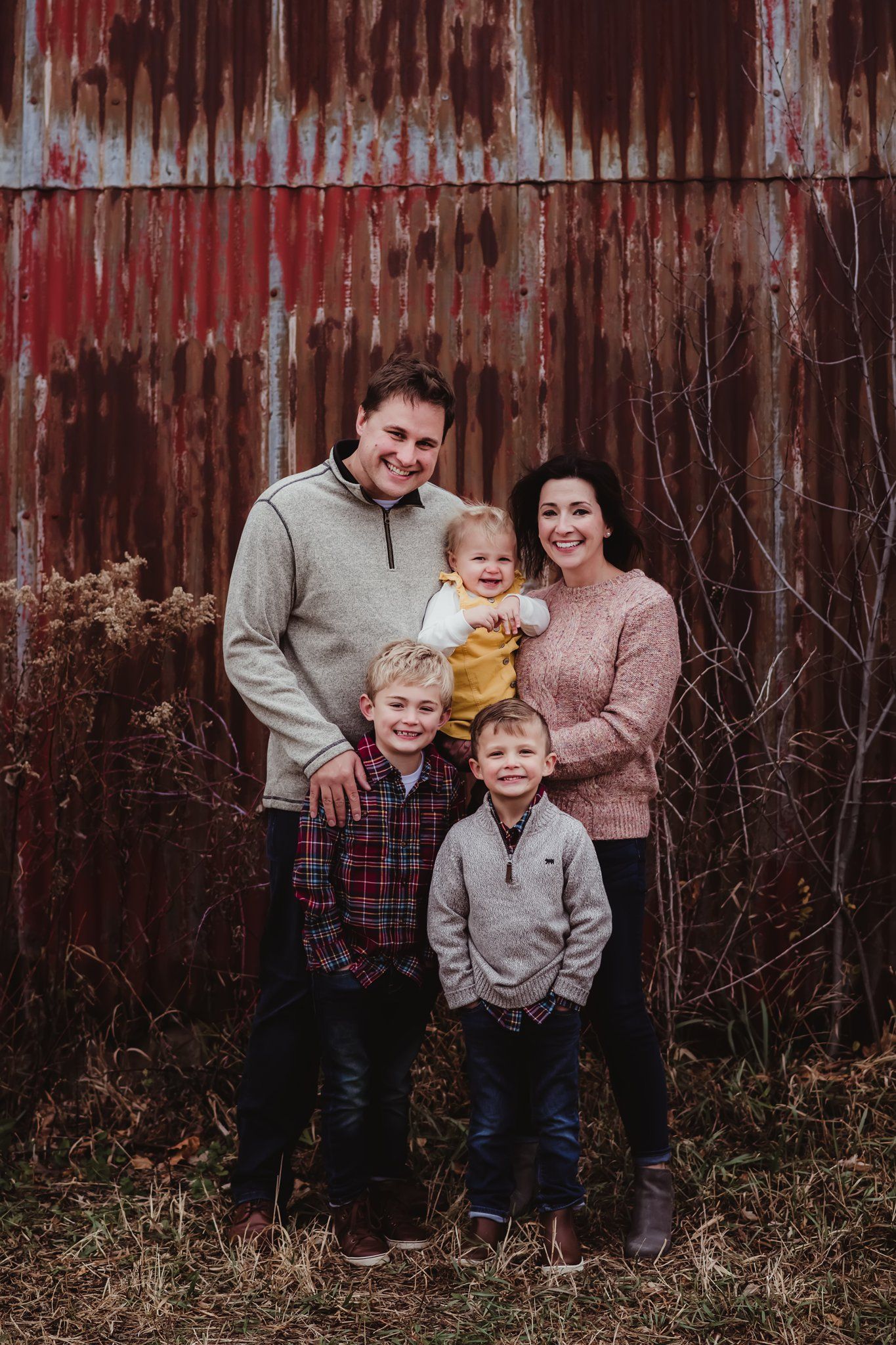 A smiling family stands together for a portrait in front of a weathered red barn wall.
