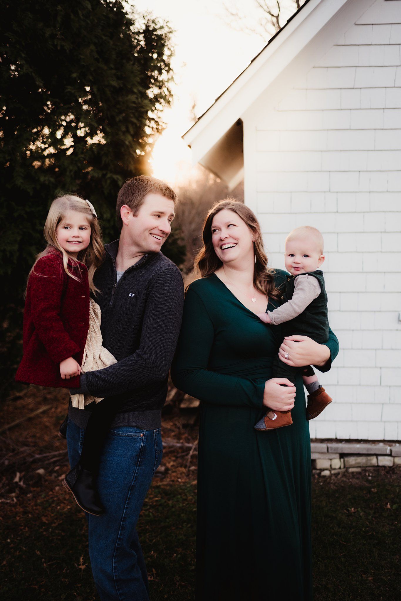 Family portrait with white house backdrop and golden sunlight peeking through trees.