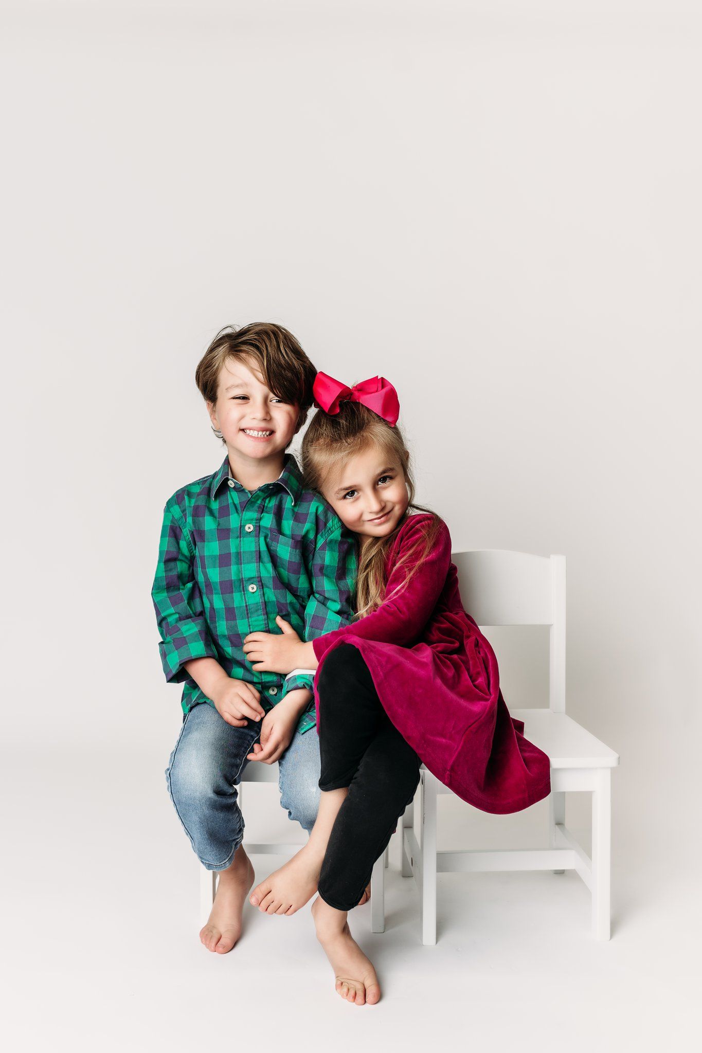 Two children in festive attire sitting together on white chair against light background.