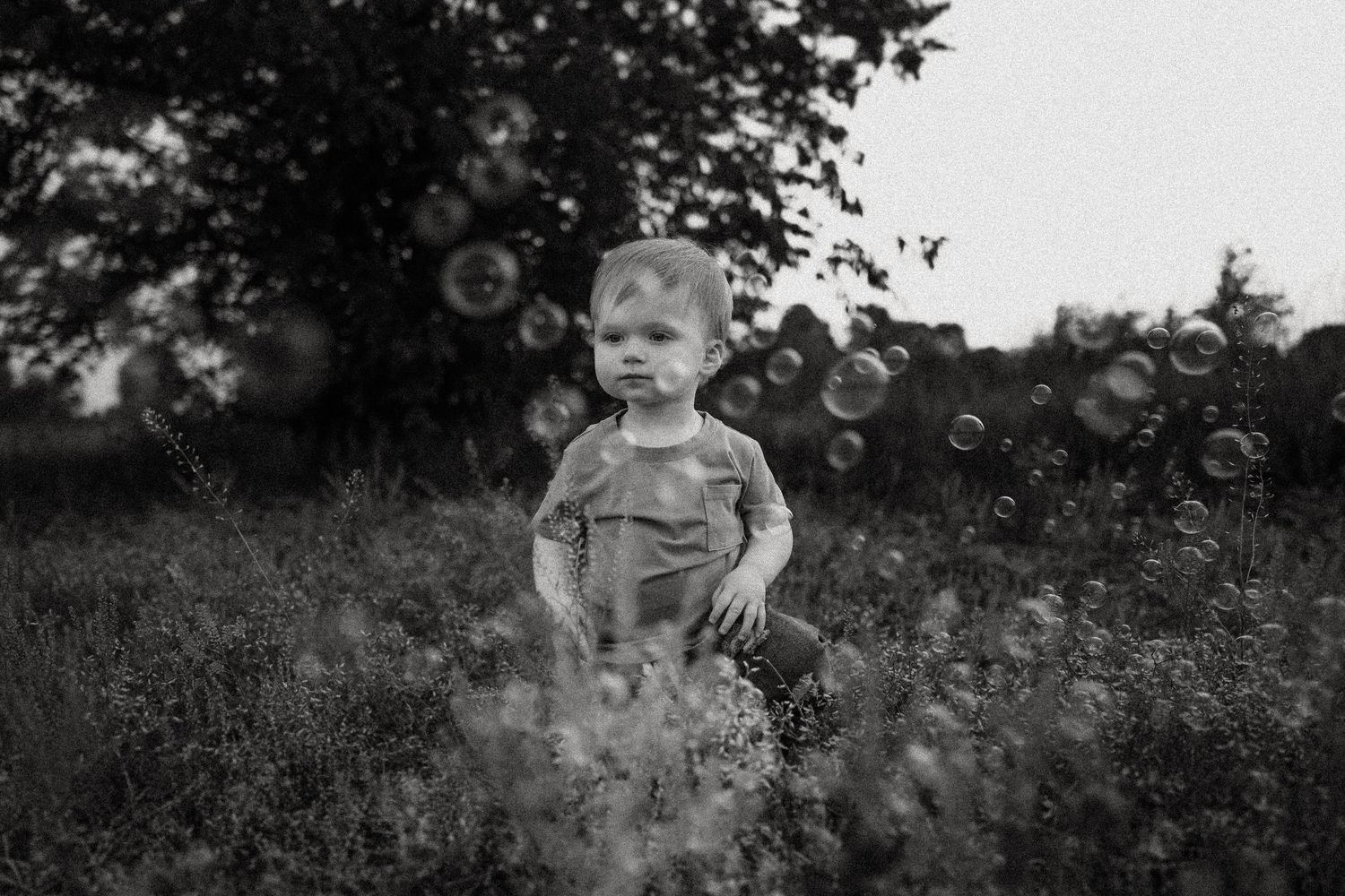 Black and white portrait of a child surrounded by floating bubbles in a field.