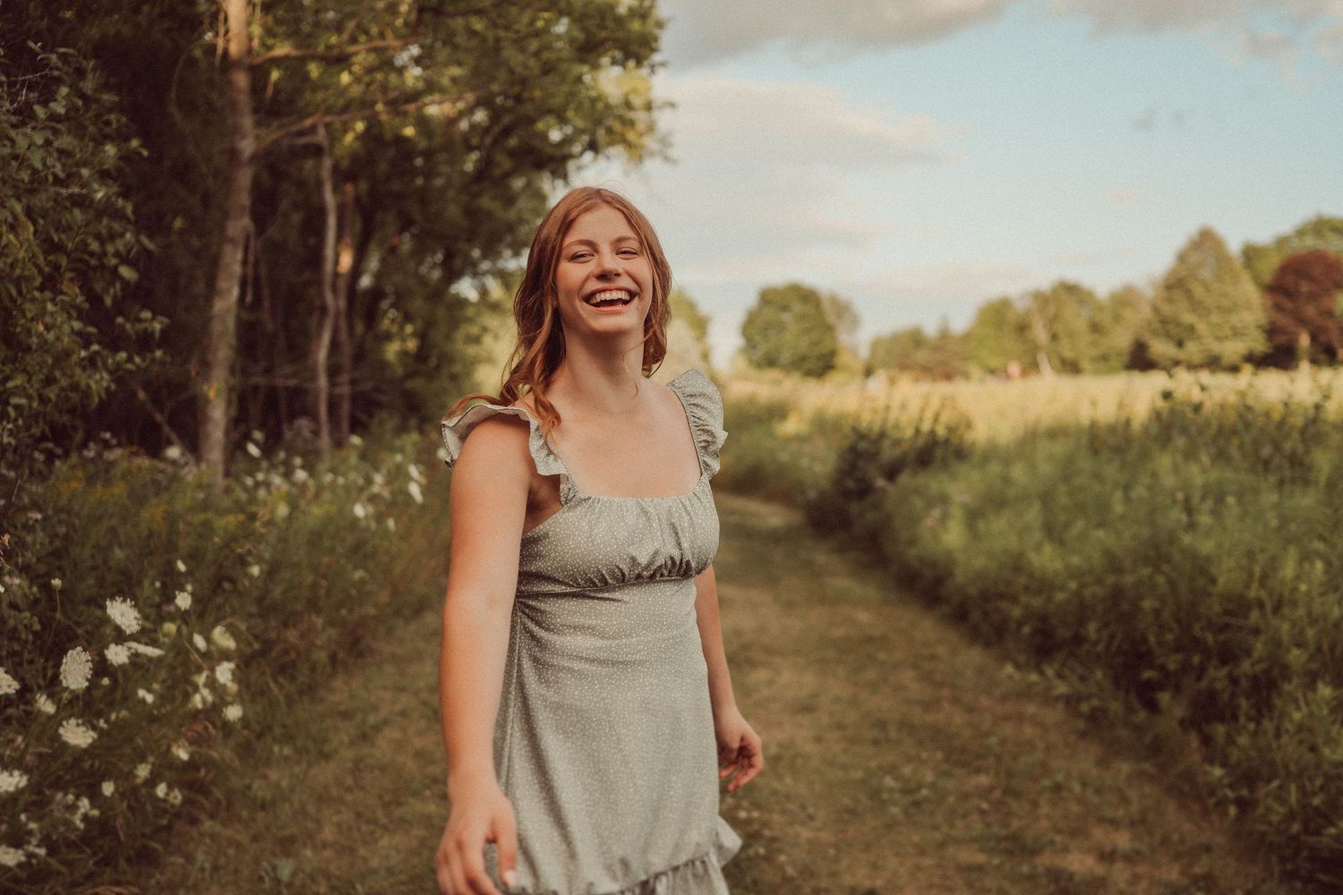 Woman in gray dress laughs joyfully while standing on a dirt path through a summer meadow.