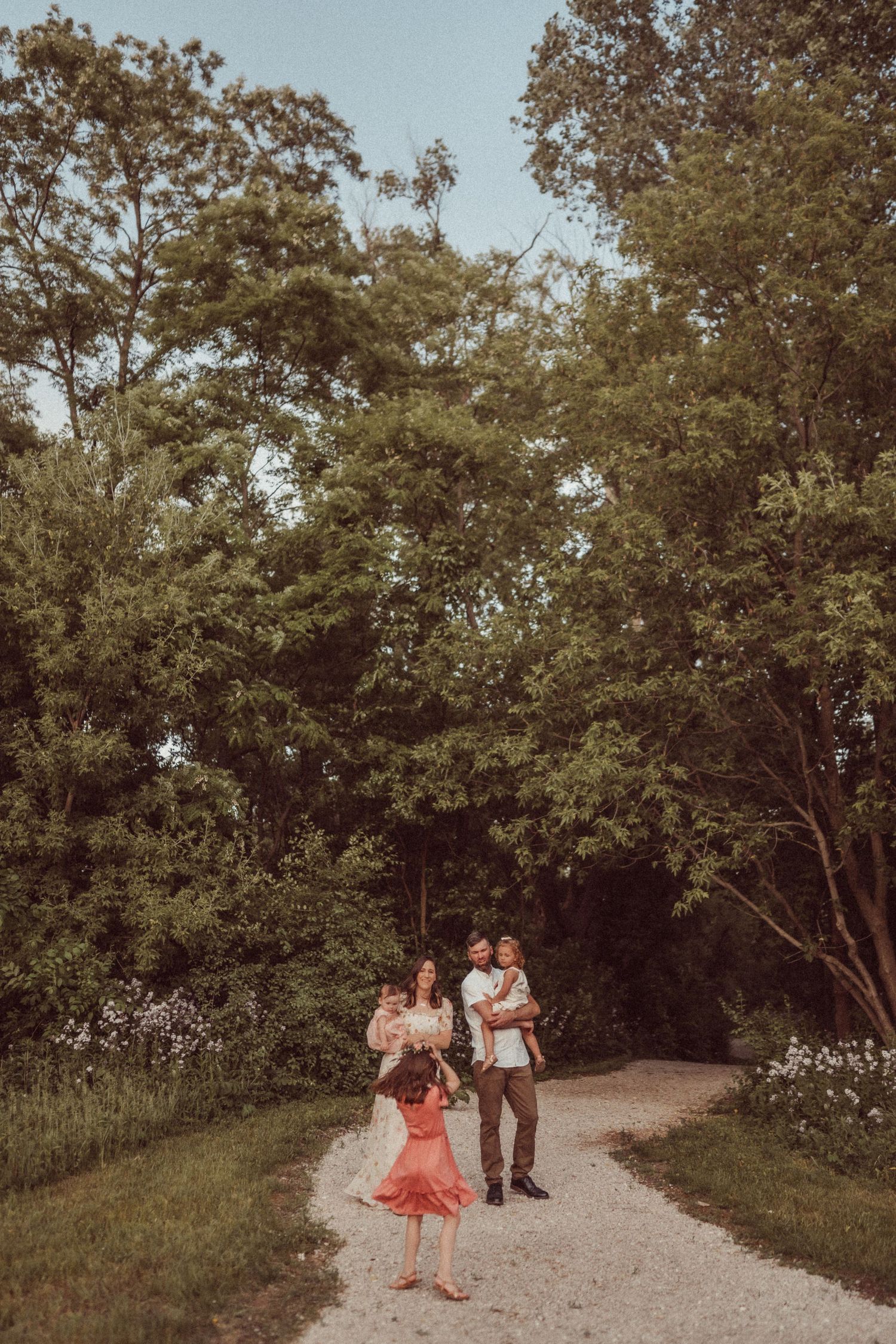 A family walks together down a winding path surrounded by tall trees and summer wildflowers.
