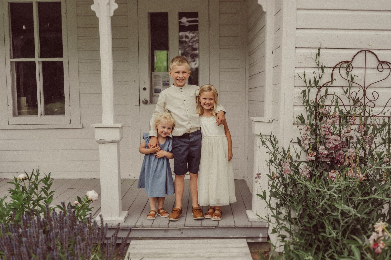 Vintage photo of children standing on white farmhouse porch with flowering garden.
