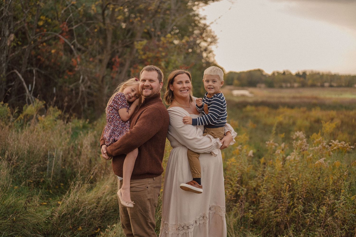 Family walking together through meadow during golden hour with autumn trees in background.