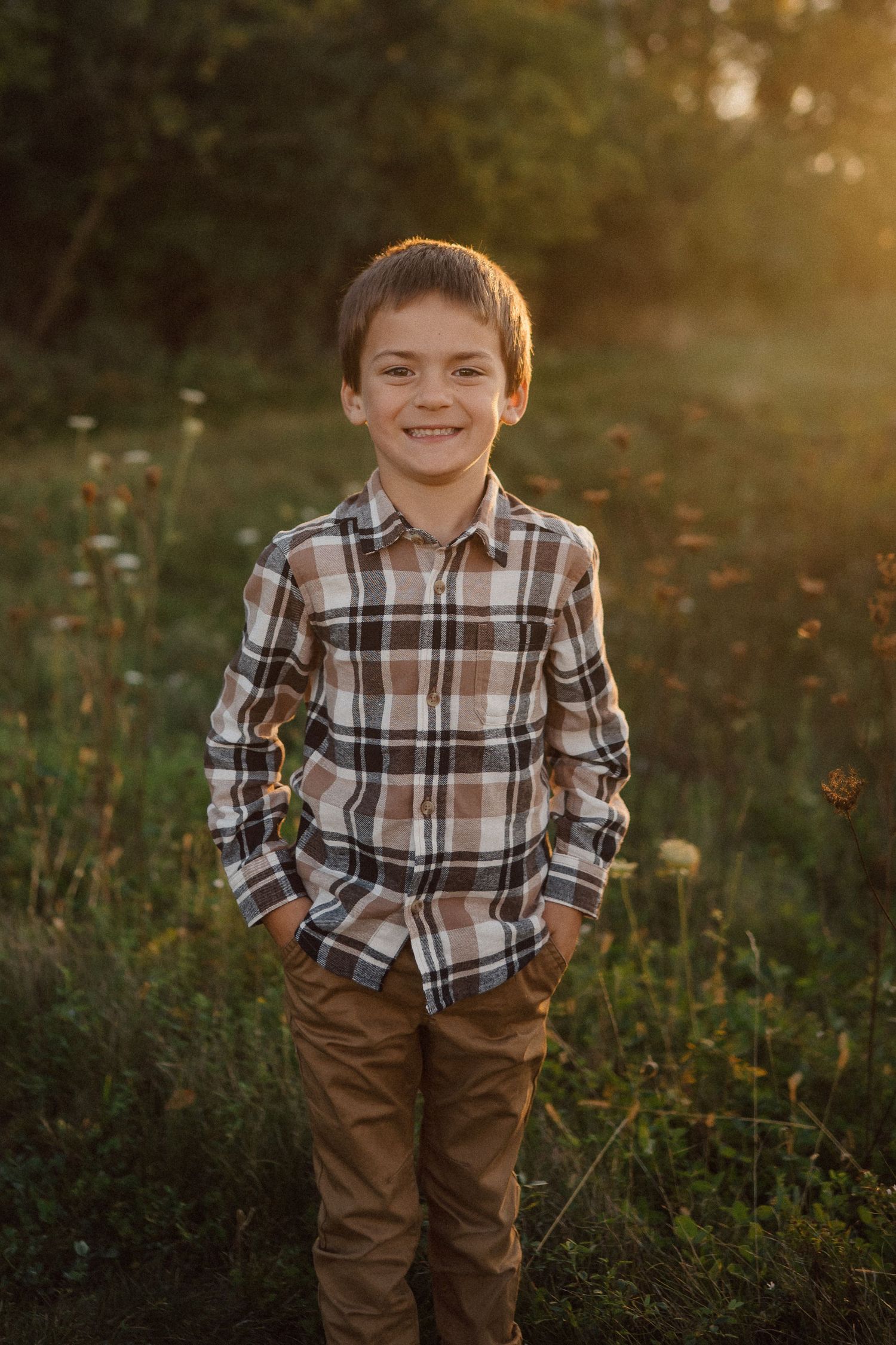 A child in a plaid shirt and brown pants poses outdoors during golden hour in a wildflower field.