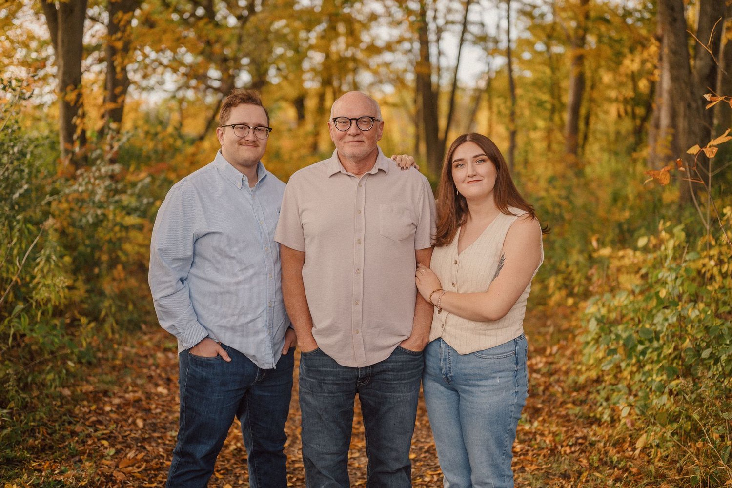 Three people pose together on a fall day surrounded by golden autumn foliage and trees in a natural outdoor setting.