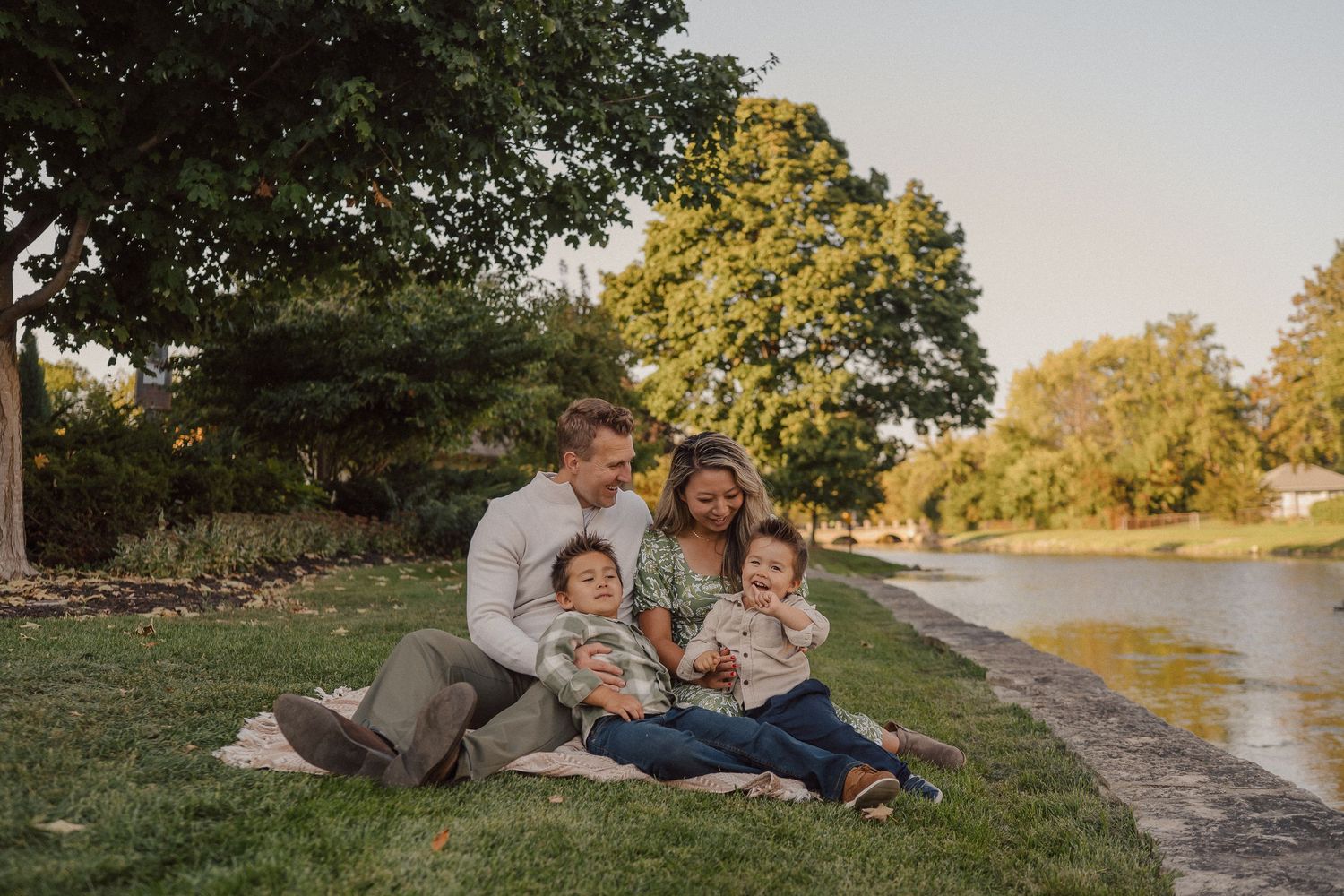 A family relaxes together outdoors during golden hour on a grassy hillside near a lake.