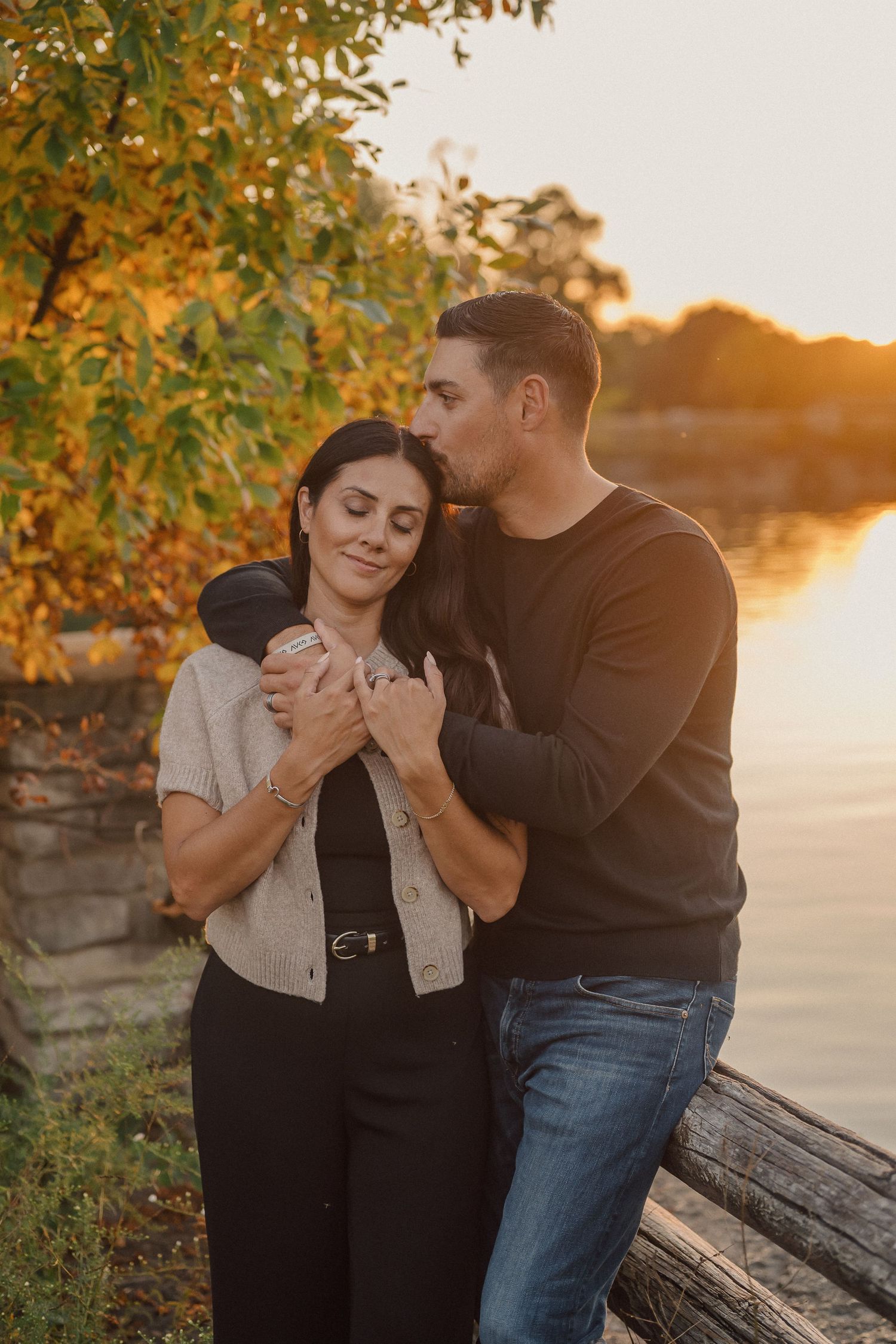 A romantic couple embraces during golden hour sunset by a lake with autumn foliage in the background.