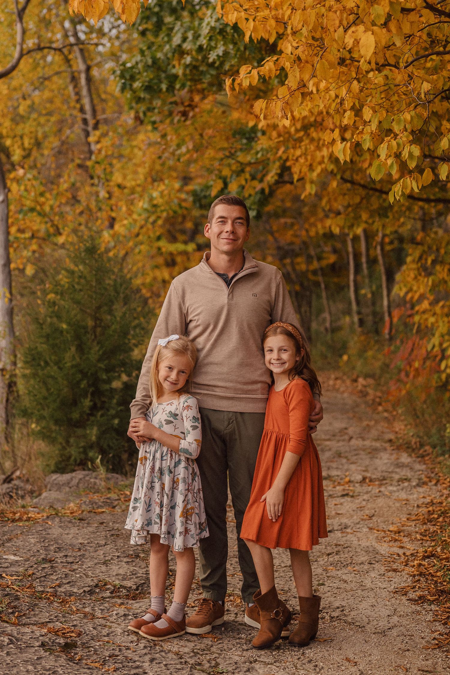 A parent and two children pose together on a wooded trail surrounded by golden autumn leaves.