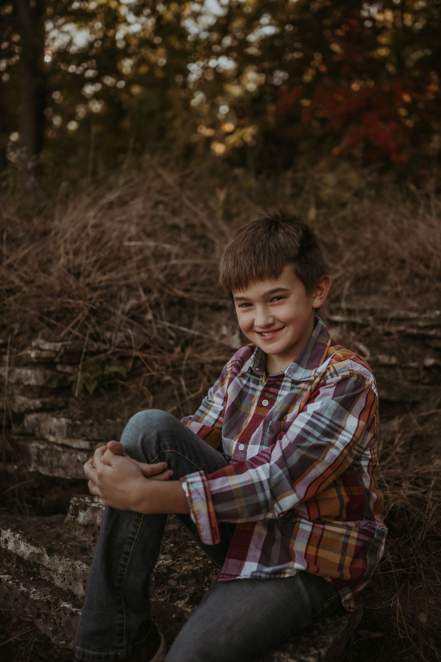Portrait in plaid shirt sitting on natural stone formation in wooded setting.