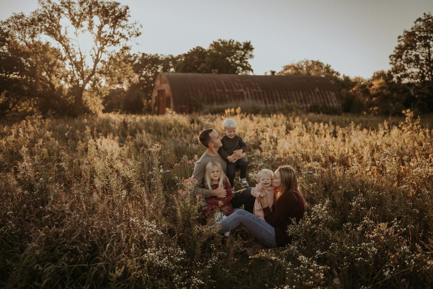 Group sitting together in wild grass field with barn in background during sunset.