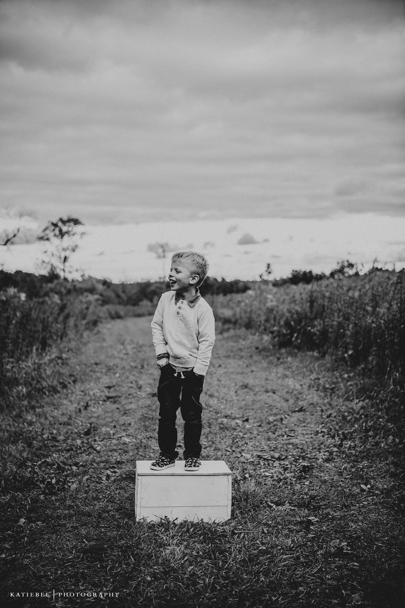 A black and white photo of a child standing on a white box on a nature path.