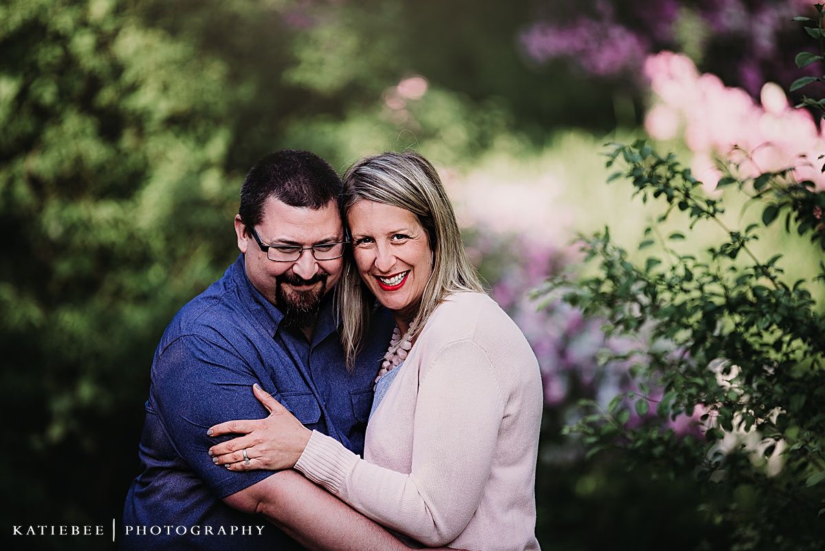 Couple sharing intimate moment among pink flowering bushes in garden setting.