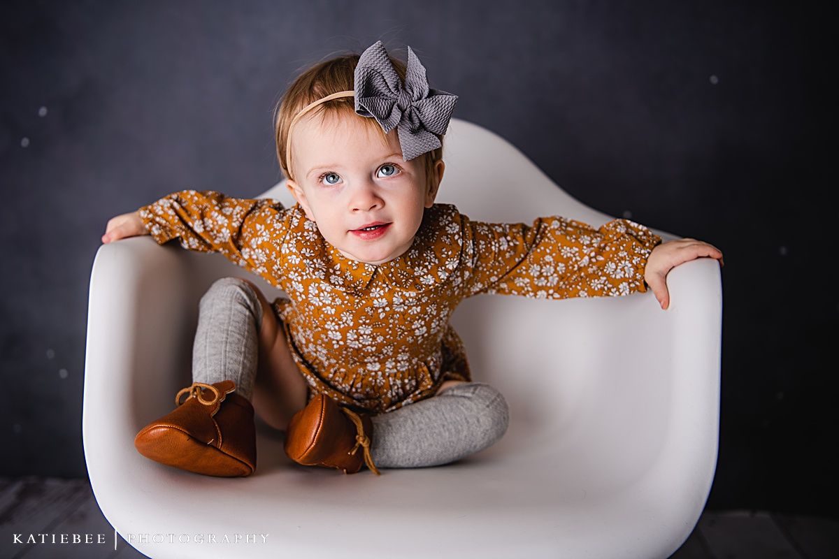 A small child wearing orange shoes and a floral dress poses playfully in a white modern chair.