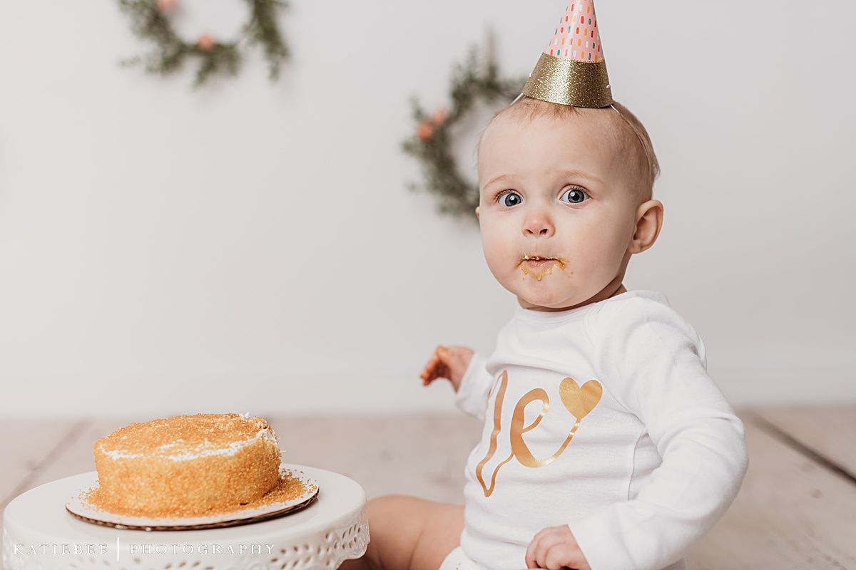 Baby wearing party hat looks at cake on white pedestal during birthday photoshoot.