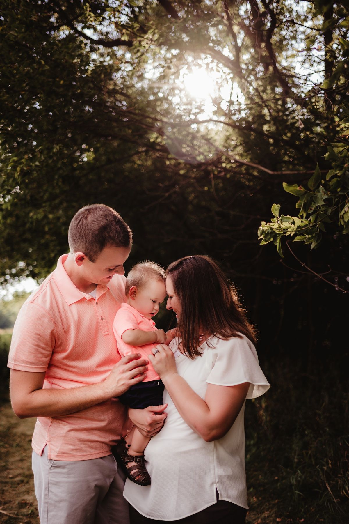 Intimate family portrait in coral and white clothing with sunburst through trees in background.