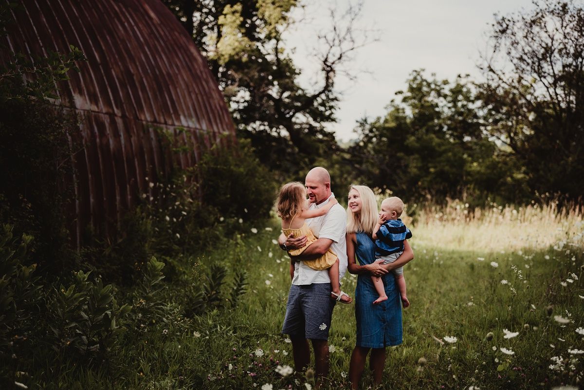 Couple holding children poses in front of weathered barn structure.