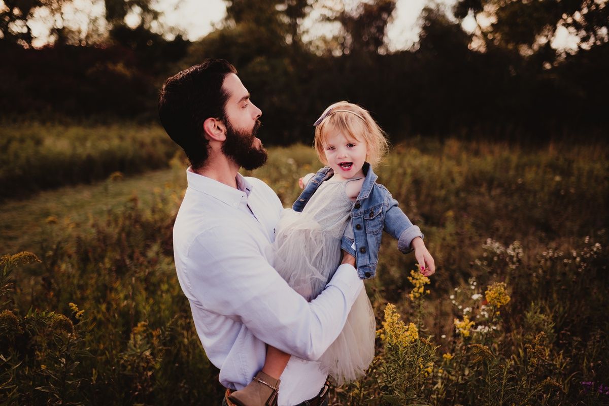 Sunset portrait of parent lifting child in jean jacket amidst golden wildflowers.