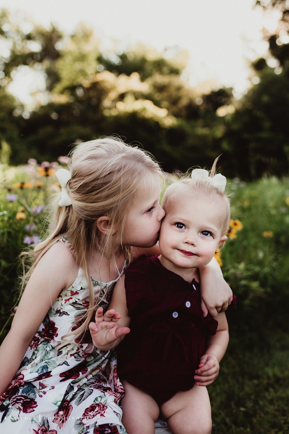 A sweet moment in nature as children share a kiss on the cheek in a garden filled with wildflowers at sunset.