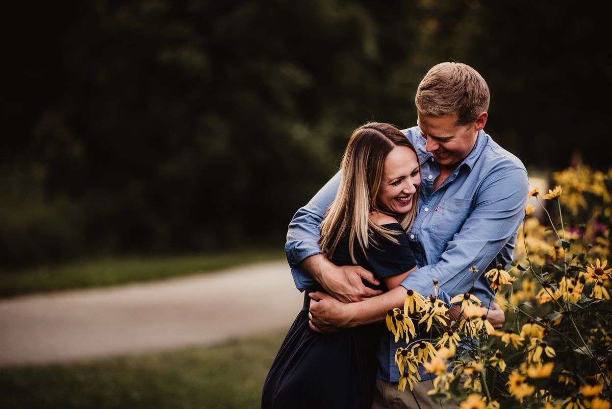 Couple laughing together while embracing near yellow flowers during sunset.