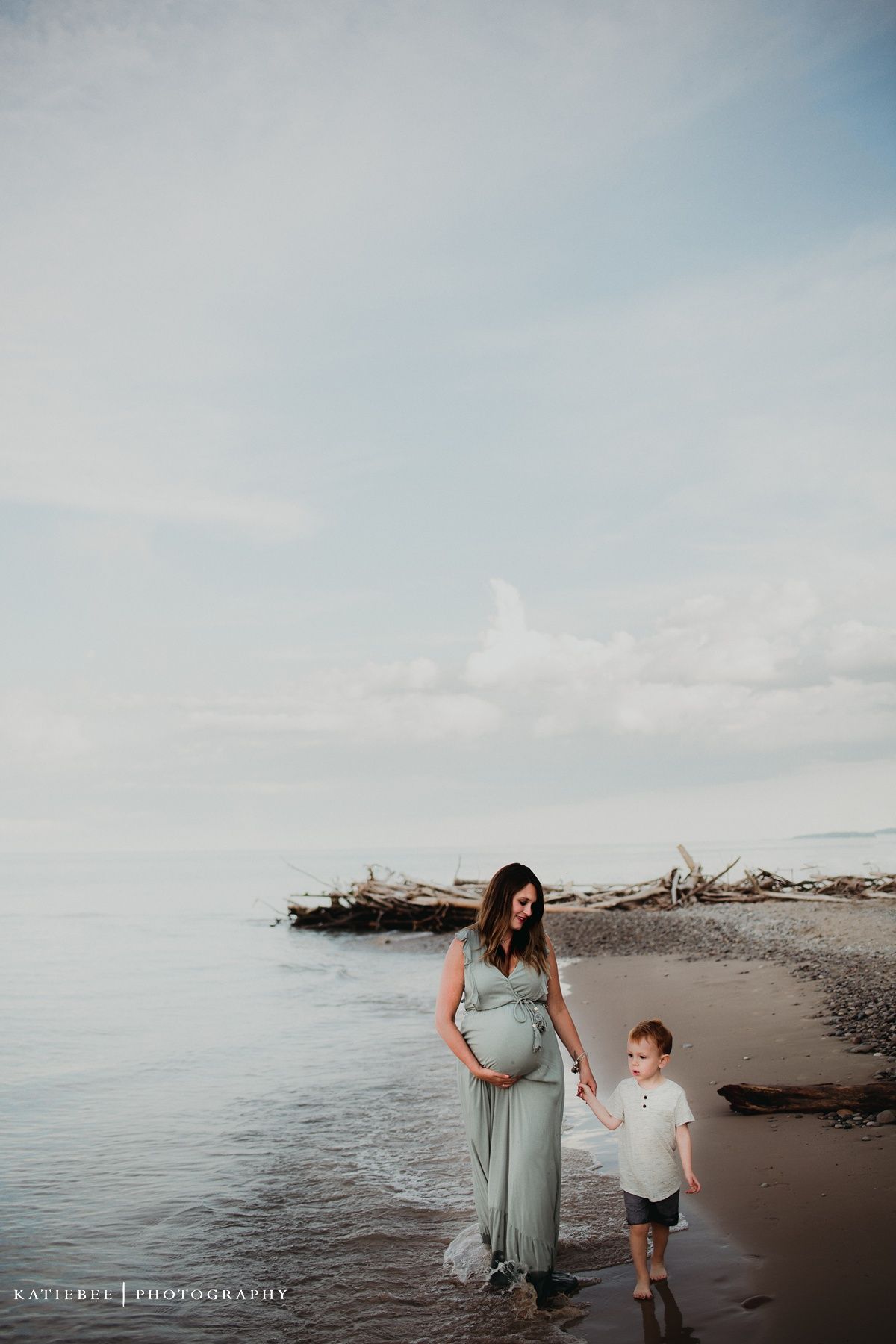 Pregnant woman in flowing sage dress walks along beach shore with young child.