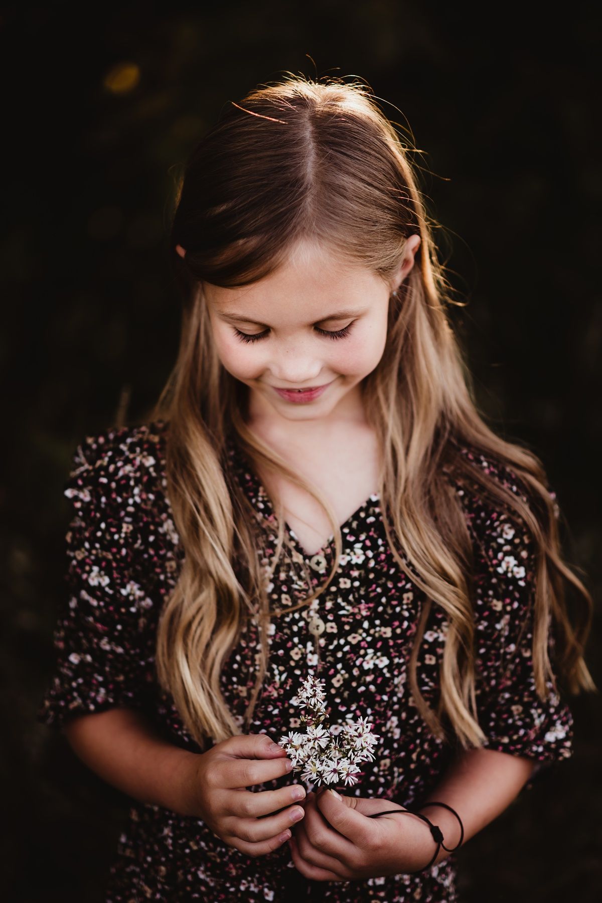 Child in floral dress looking down with soft smile in outdoor portrait.