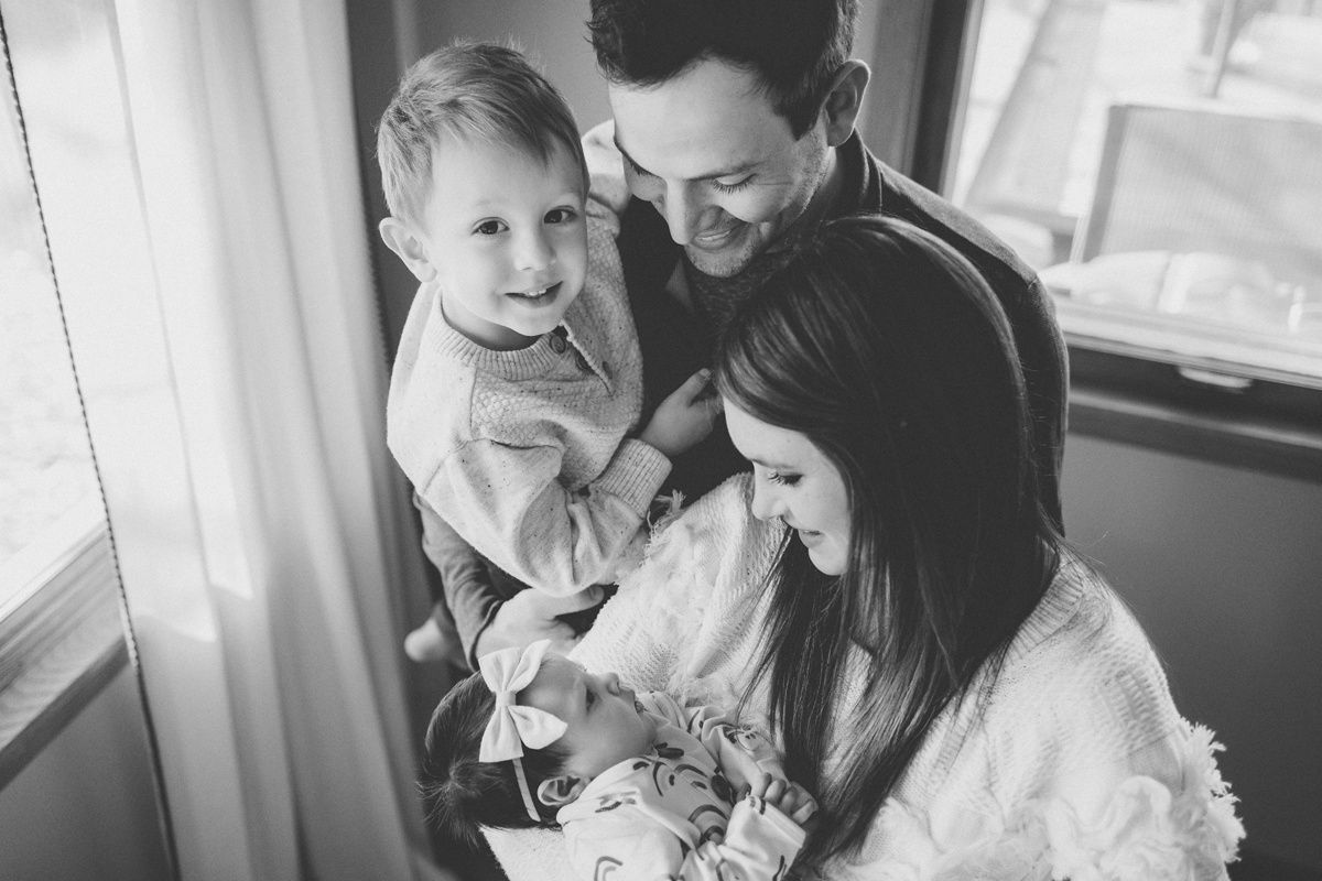 Family portrait in natural light with a toddler and newborn by the window.
