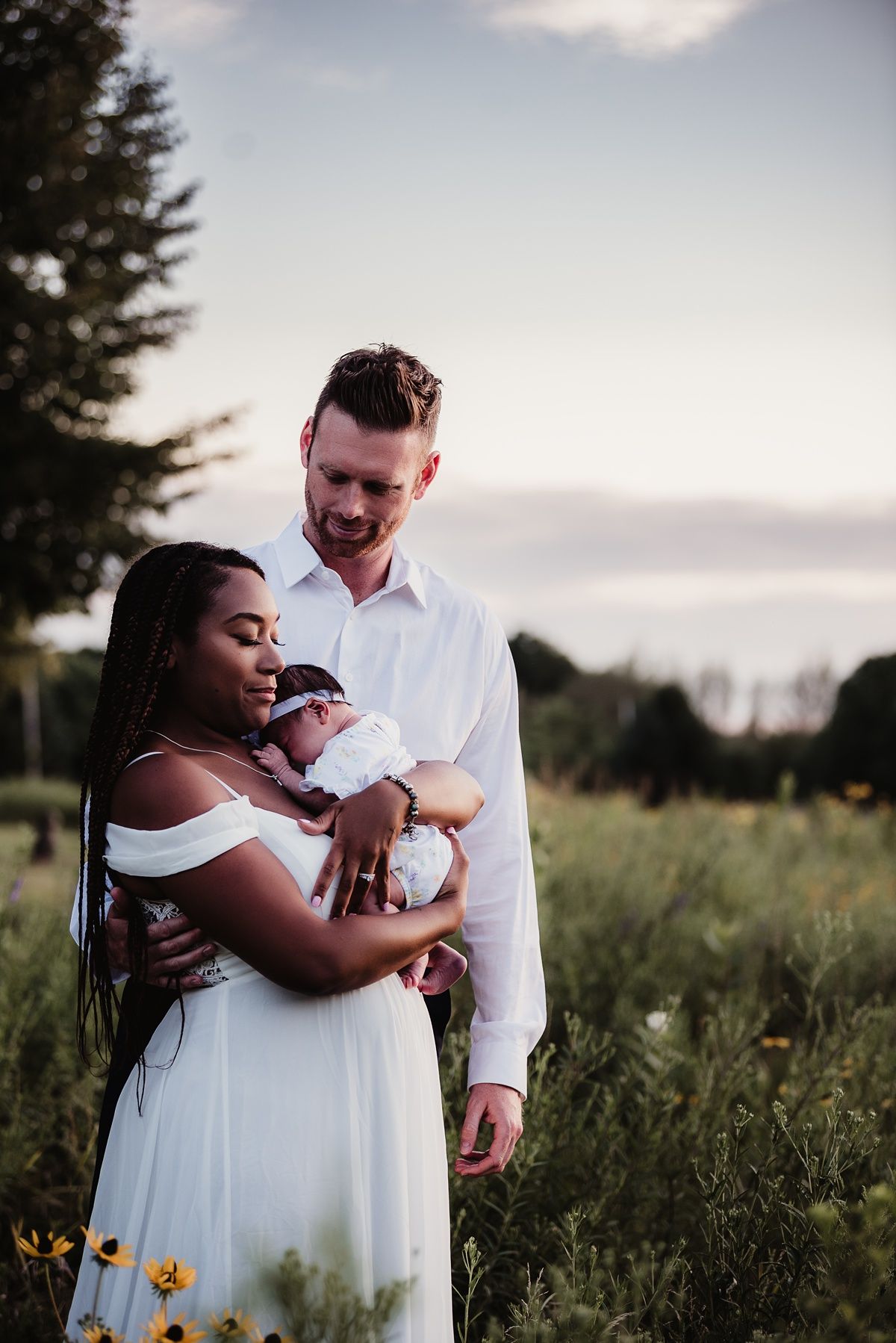 A couple in white clothing shares an intimate moment with their baby in a wildflower field at sunset.