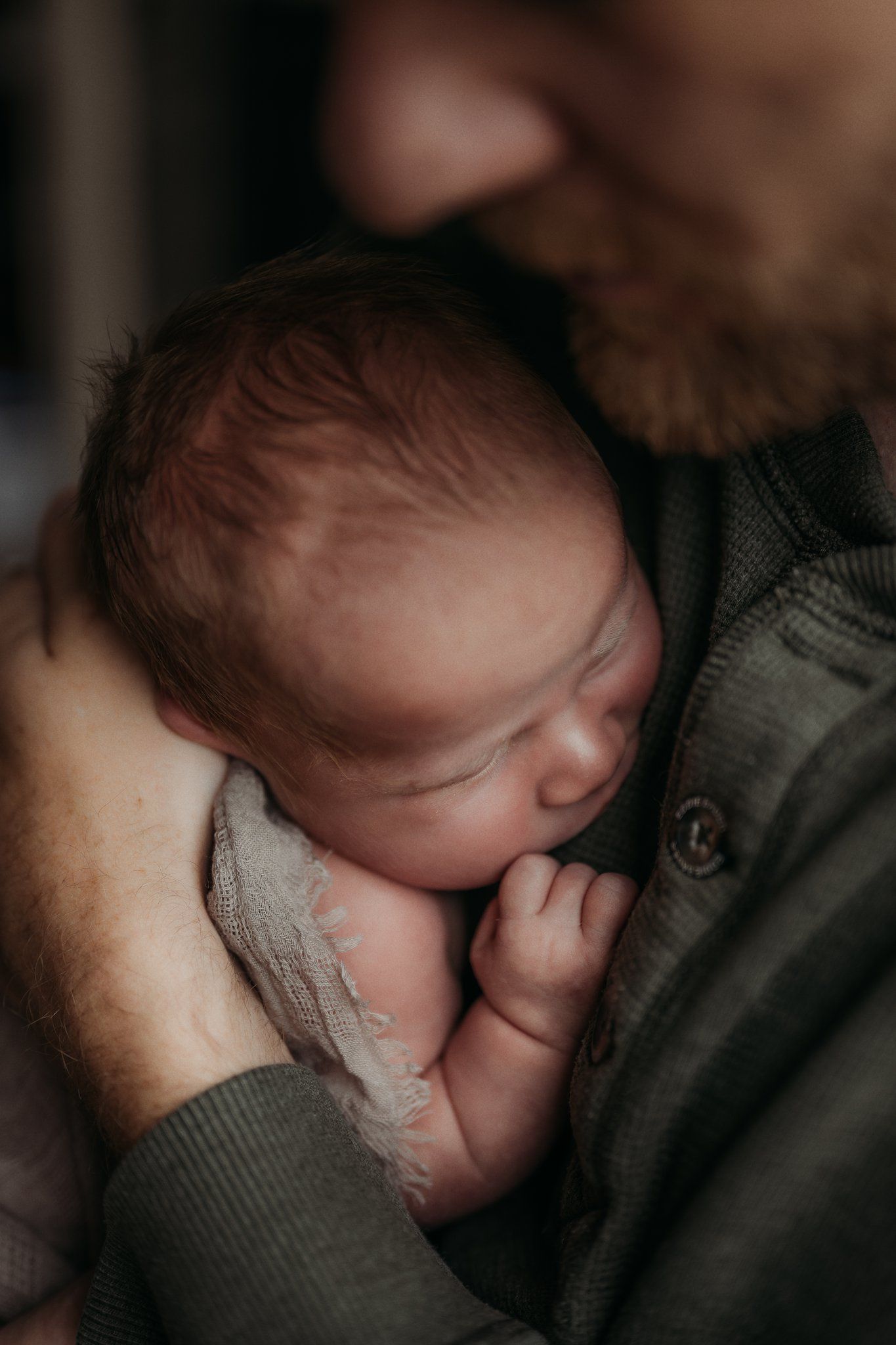 Sleeping newborn nestled against parent wearing gray sweater in dim light.