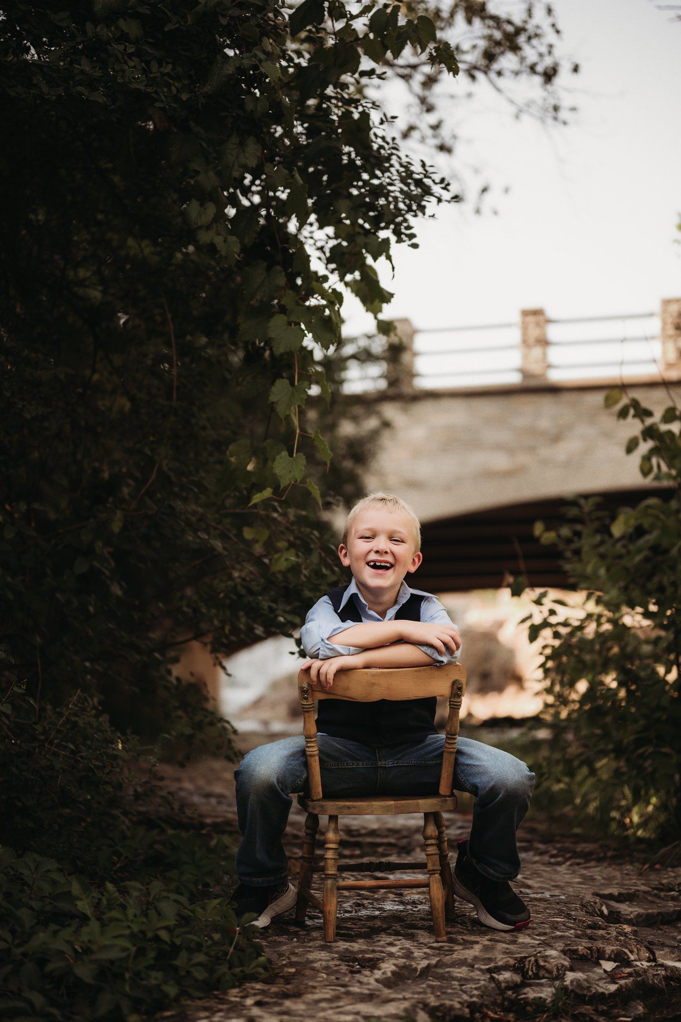 Child sitting on wooden chair outdoors under bridge with joyful expression.