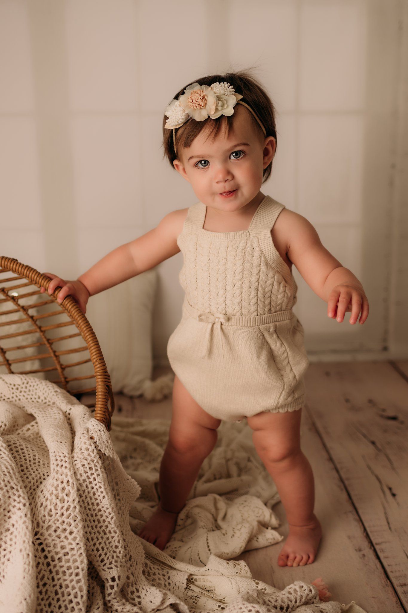 Baby in cream romper standing by rattan chair with knit blankets and greenery.