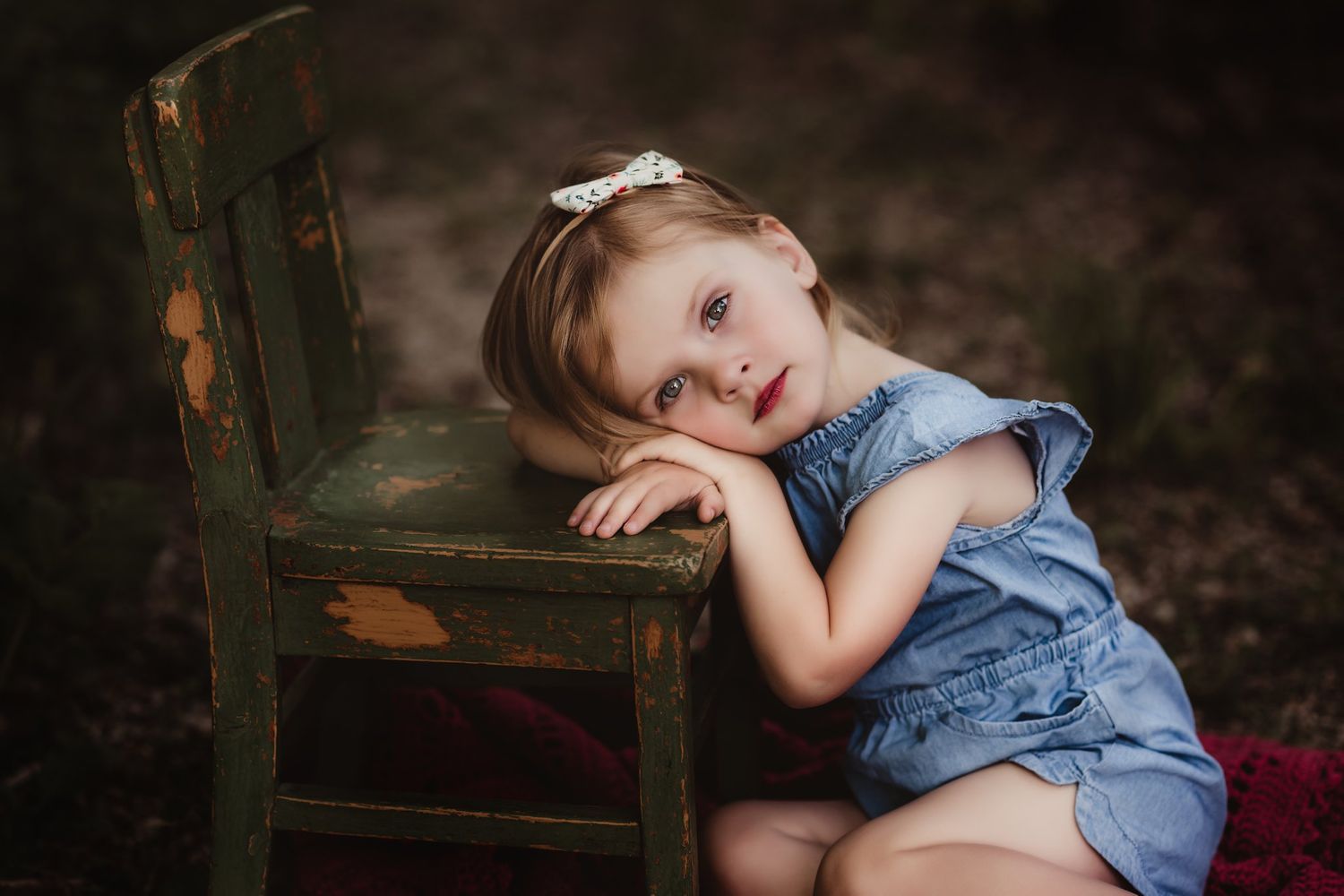 Child in blue outfit resting head on vintage wooden chair in outdoor setting.