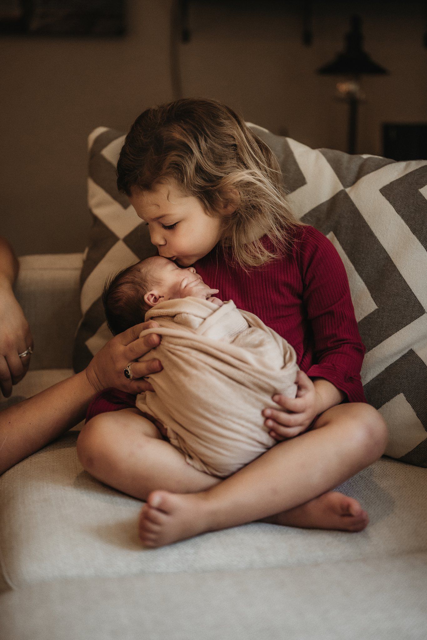 Young girl in red sweater gently kisses newborn sibling on couch with geometric pillows.