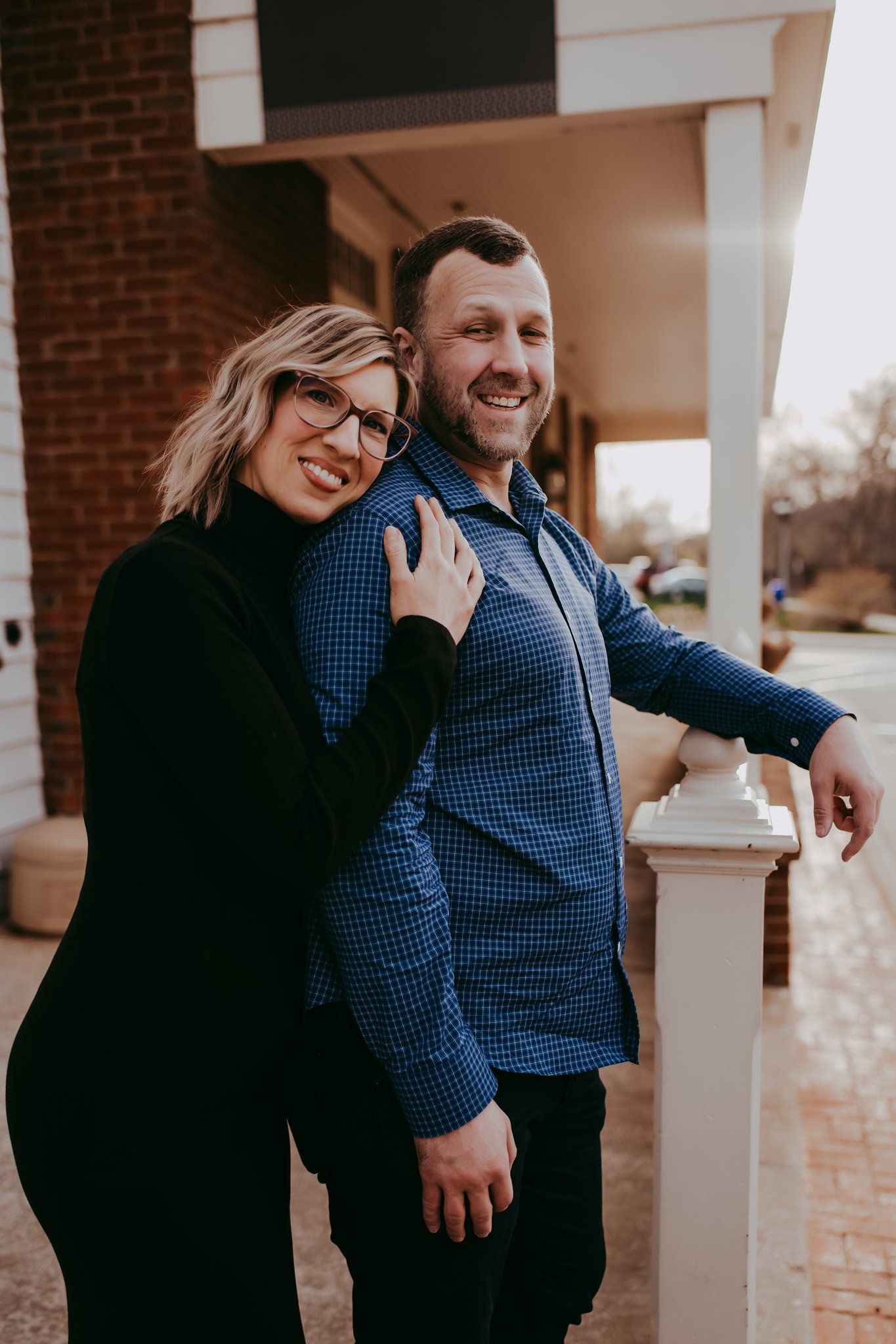 A couple in casual attire share an affectionate moment against a brick building backdrop.