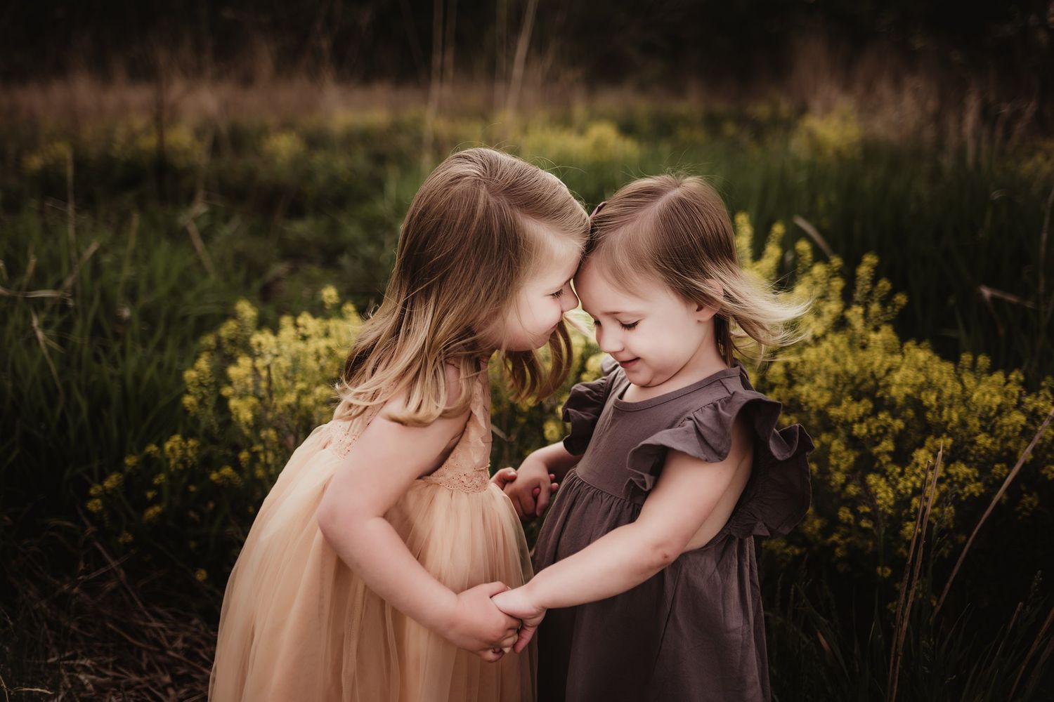 Two children in flowing dresses share intimate moment among yellow flowers at sunset.