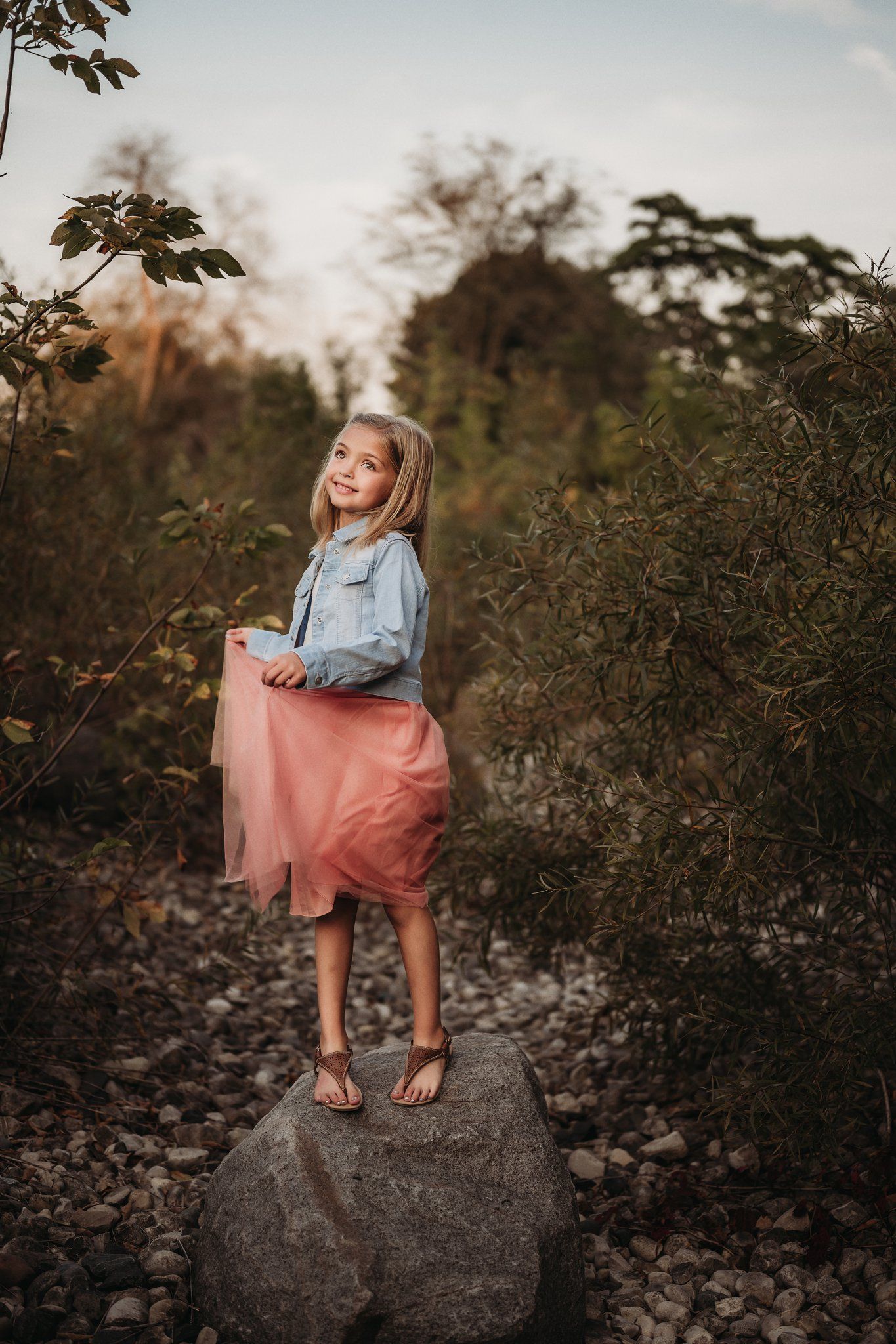 A child in denim and pink tulle stands confidently on a large rock during sunset.