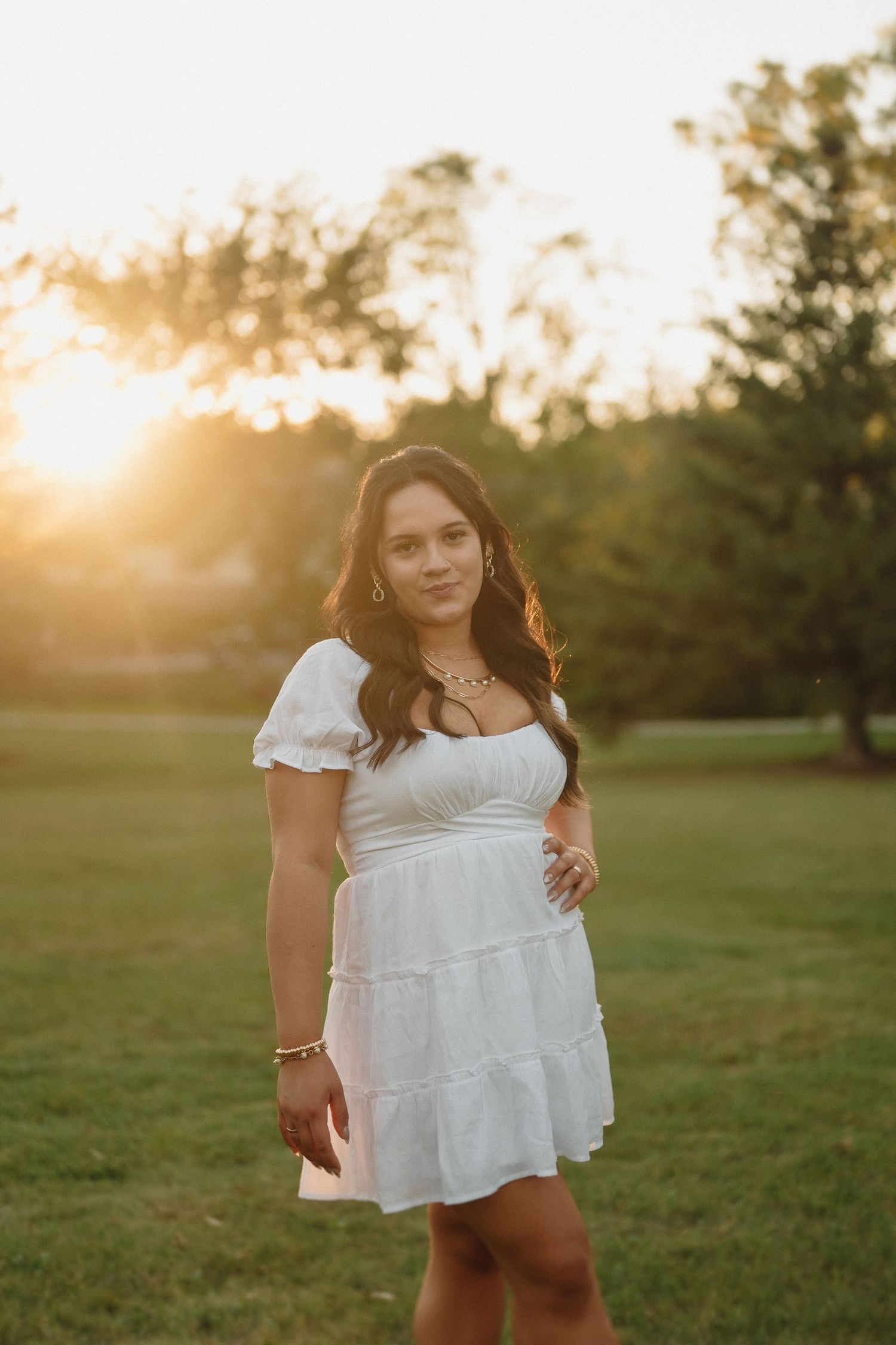 Woman in white summer dress poses in field during golden hour sunset with glowing backlight.