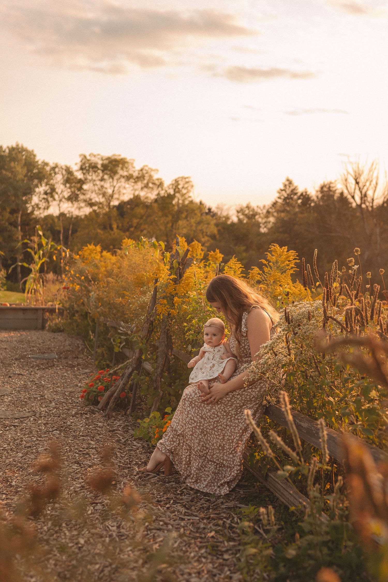 Mother and baby sit on garden path during golden hour surrounded by wildflowers.