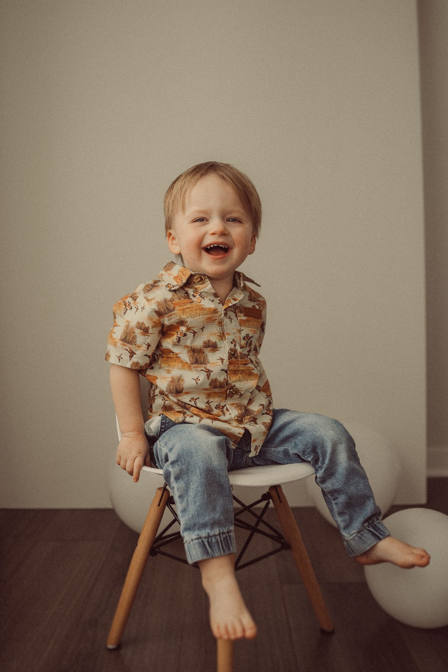 Joyful toddler in floral shirt sitting on modern white chair against neutral backdrop.