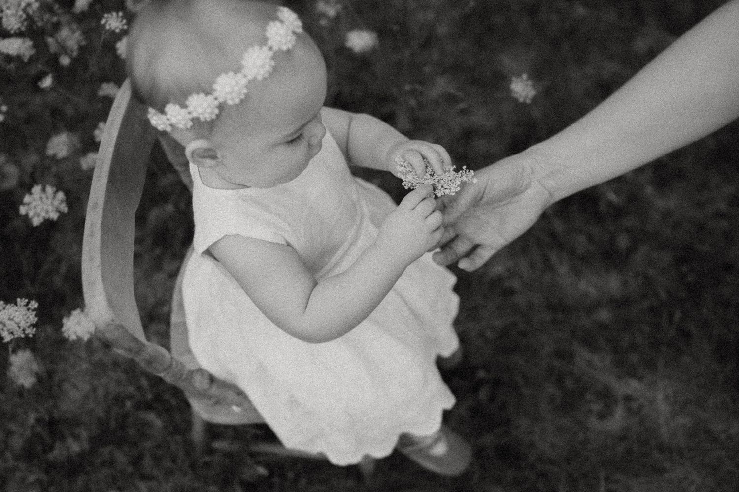 Black and white image of baby in white dress holding an adult's hand outdoors.