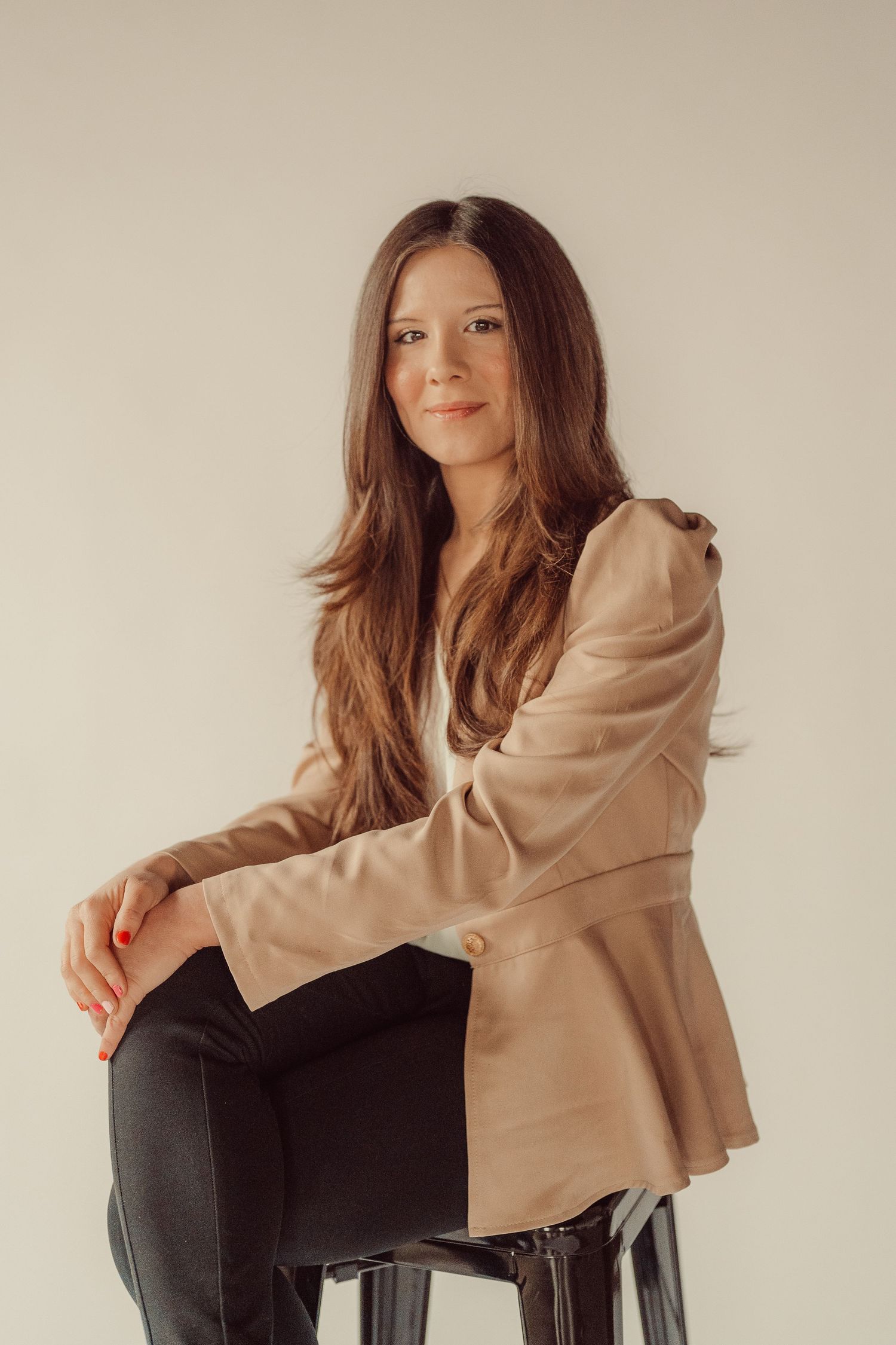 Stylish woman in tan blazer sitting on stool during professional photoshoot.