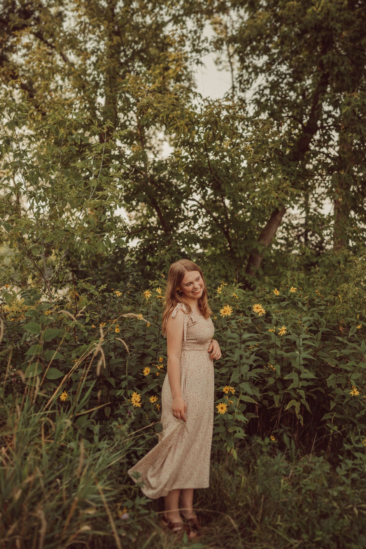 A vintage-style portrait in a long dress standing among tall sunflowers and trees.