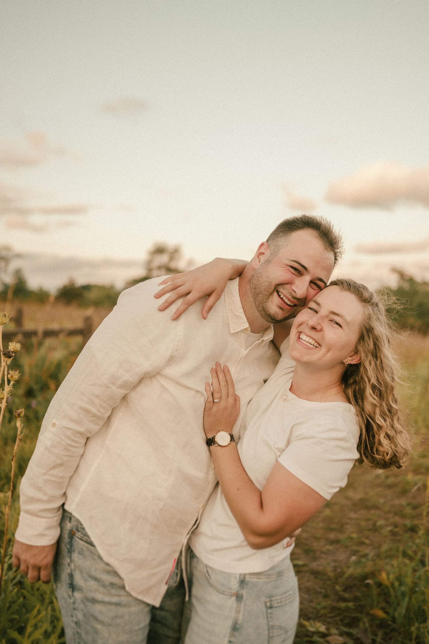 A candid moment of a couple laughing together in casual attire against a sunset sky.
