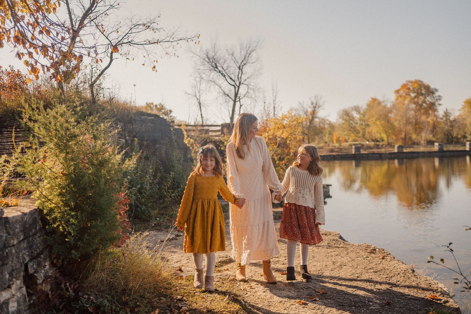 Family wearing vintage style dresses walks along autumn riverside path at golden hour.