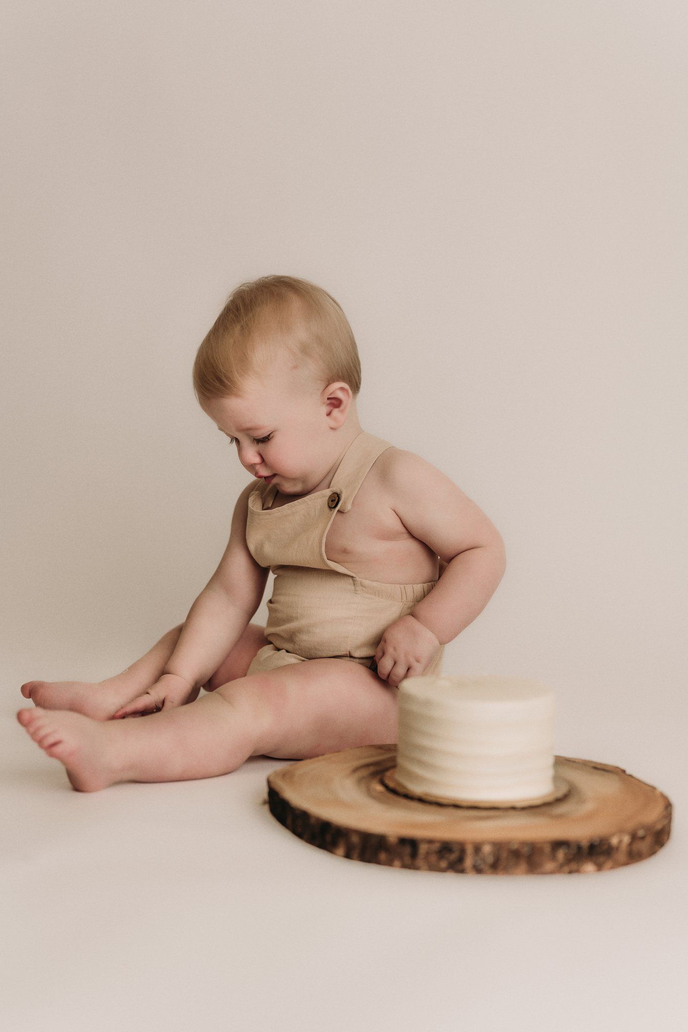 Baby in beige romper reaches down while sitting next to white cake on wood slice.