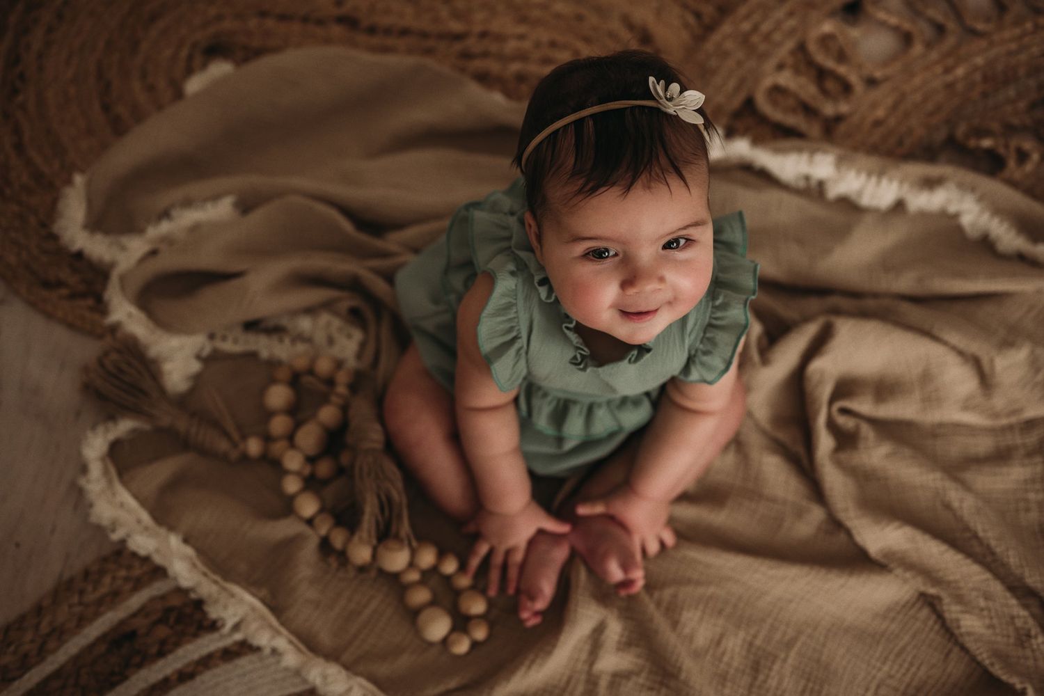 A baby in green ruffled dress poses on beige blankets with wooden bead decorations.
