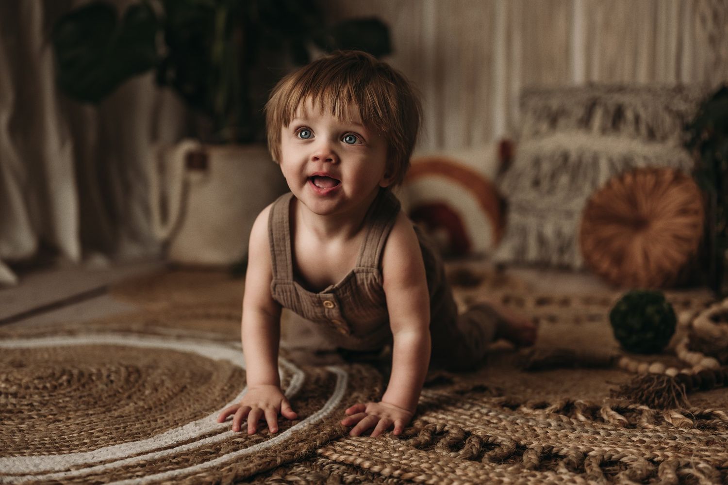 A baby in brown attire explores a textured carpet in a room with macrame wall decor.
