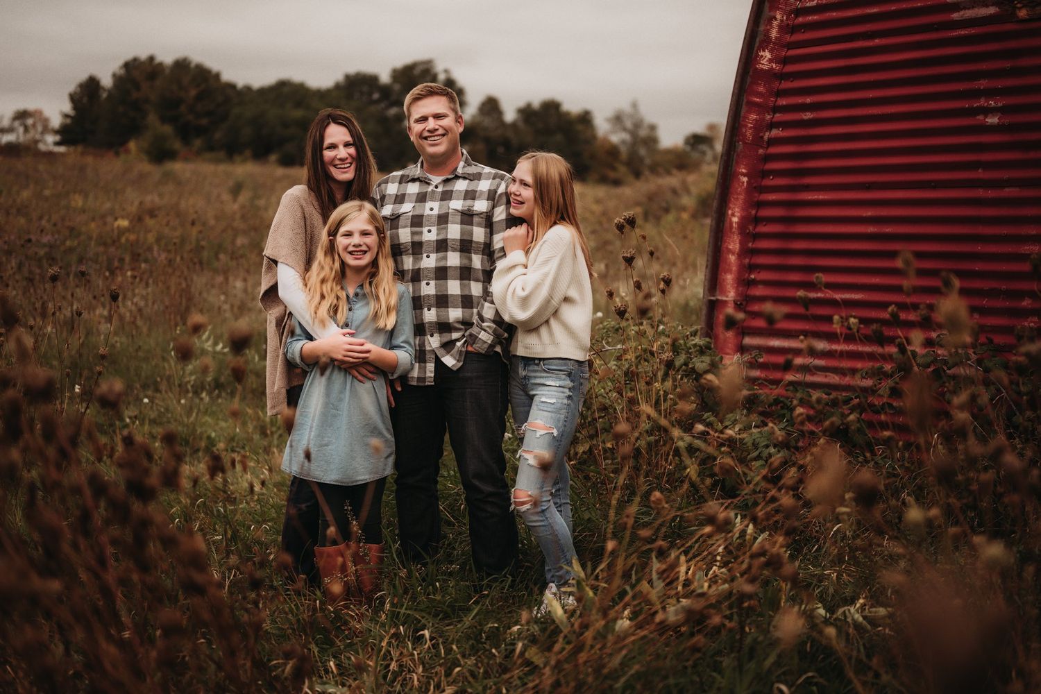 Family poses together in casual wear near red barn in autumn field.