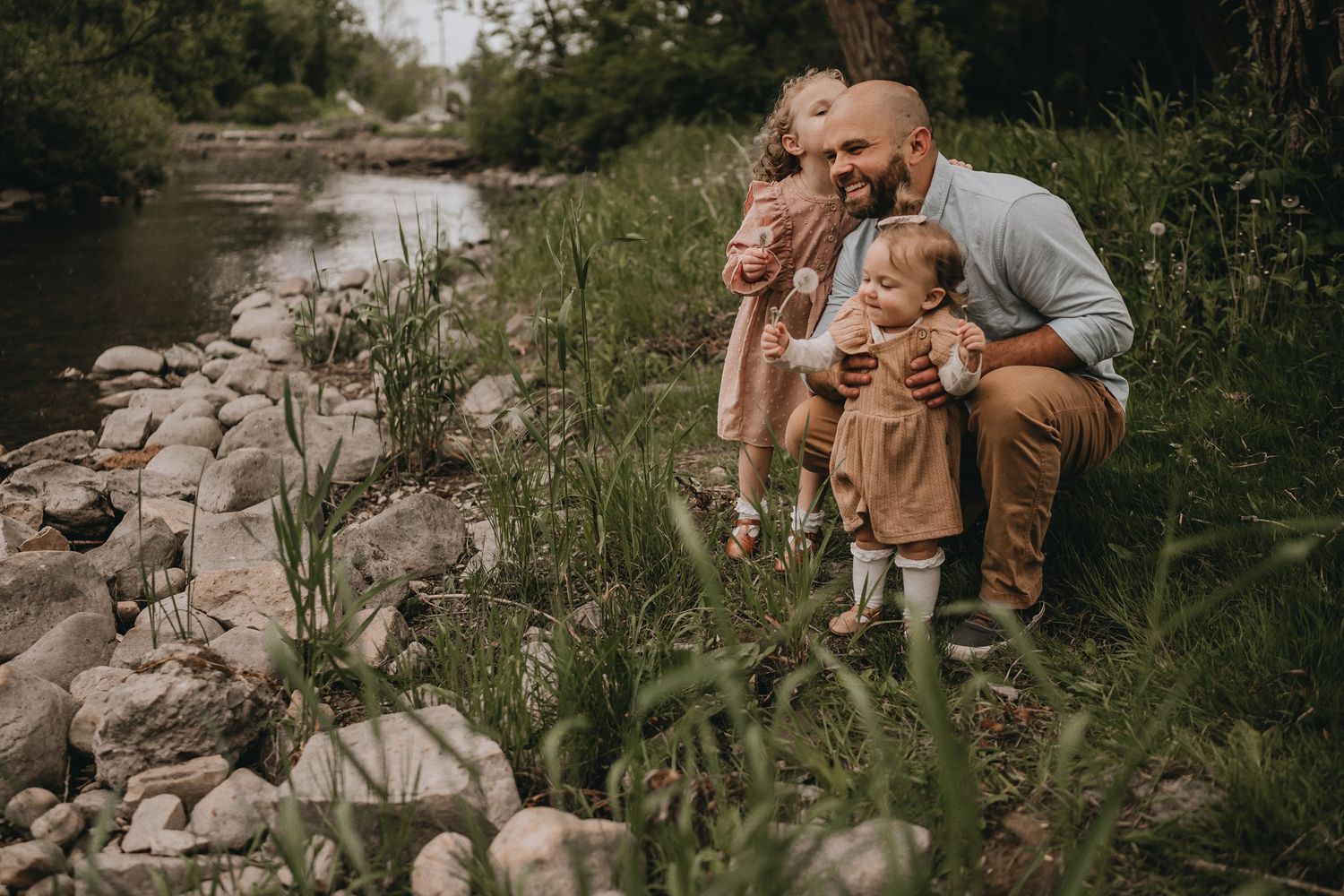 A family shares a tender moment together while sitting on rocky riverbank surrounded by grass.