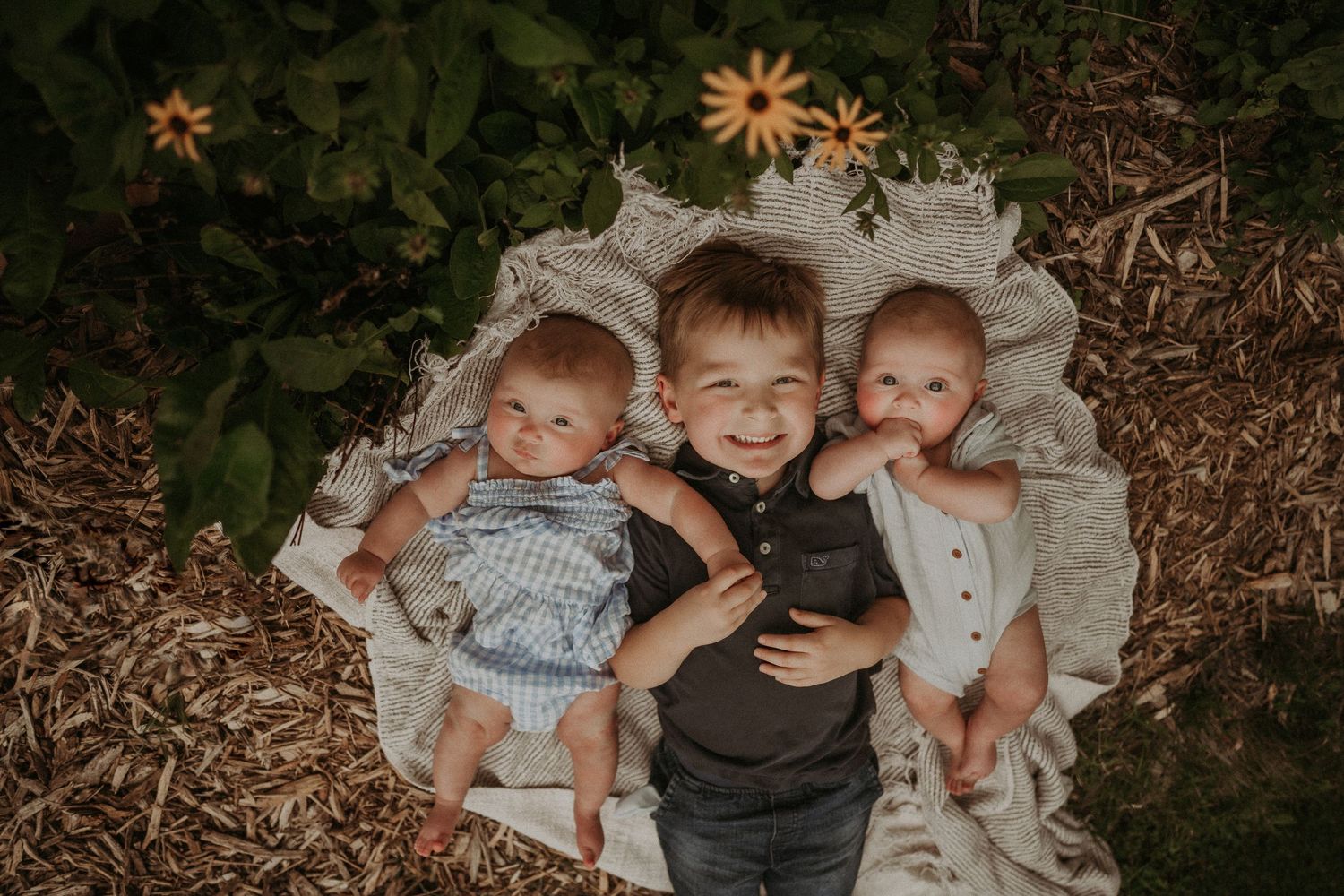 Three children laying on blanket together in garden with flowers nearby.